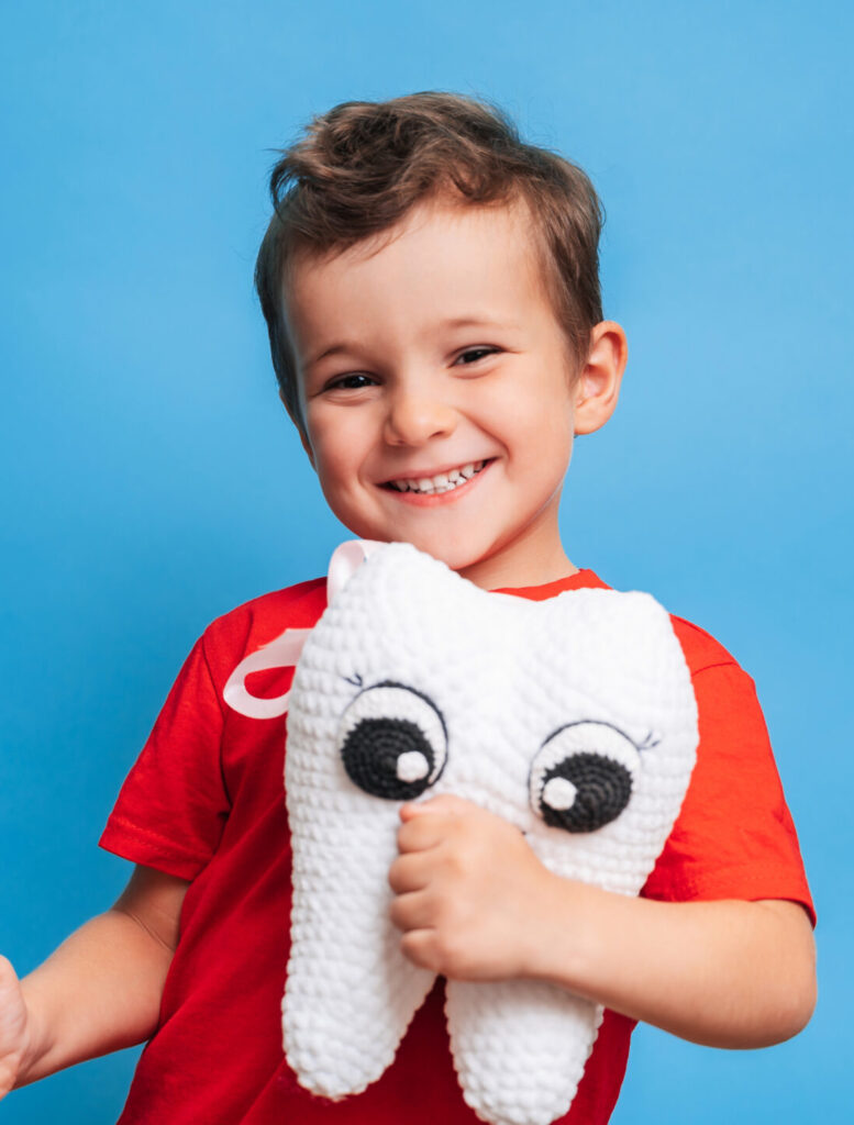 A smiling boy with healthy teeth holds a plush tooth and a toothbrush on a blue isolated background. Oral hygiene. Pediatric dentistry. Prevention of caries. A place for your text