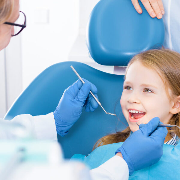 Little cute smiling girl is sitting in dental chair and opening mouth in clinic, office. Doctor is preparing for examination of child teeth with tools, instruments. Visiting dentist with children.