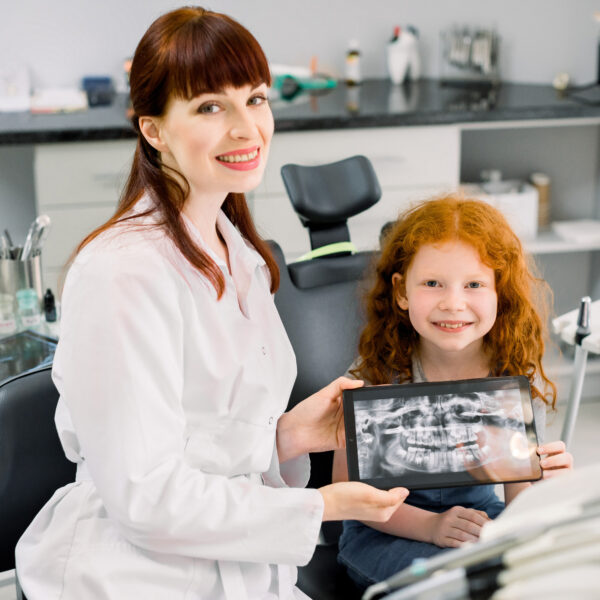Little smiling girl with red curly hair sitting on chair and looking at camera, while holding x-ray scan image of her teeth on digital tablet together with her cheerful female dentist
