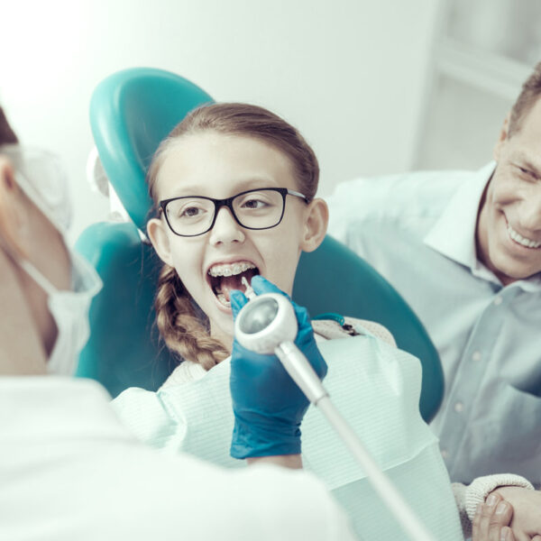 Funny procedure. Pretty little smiling girl looking happy during the dental procedure while the dentist holding a special professional engine