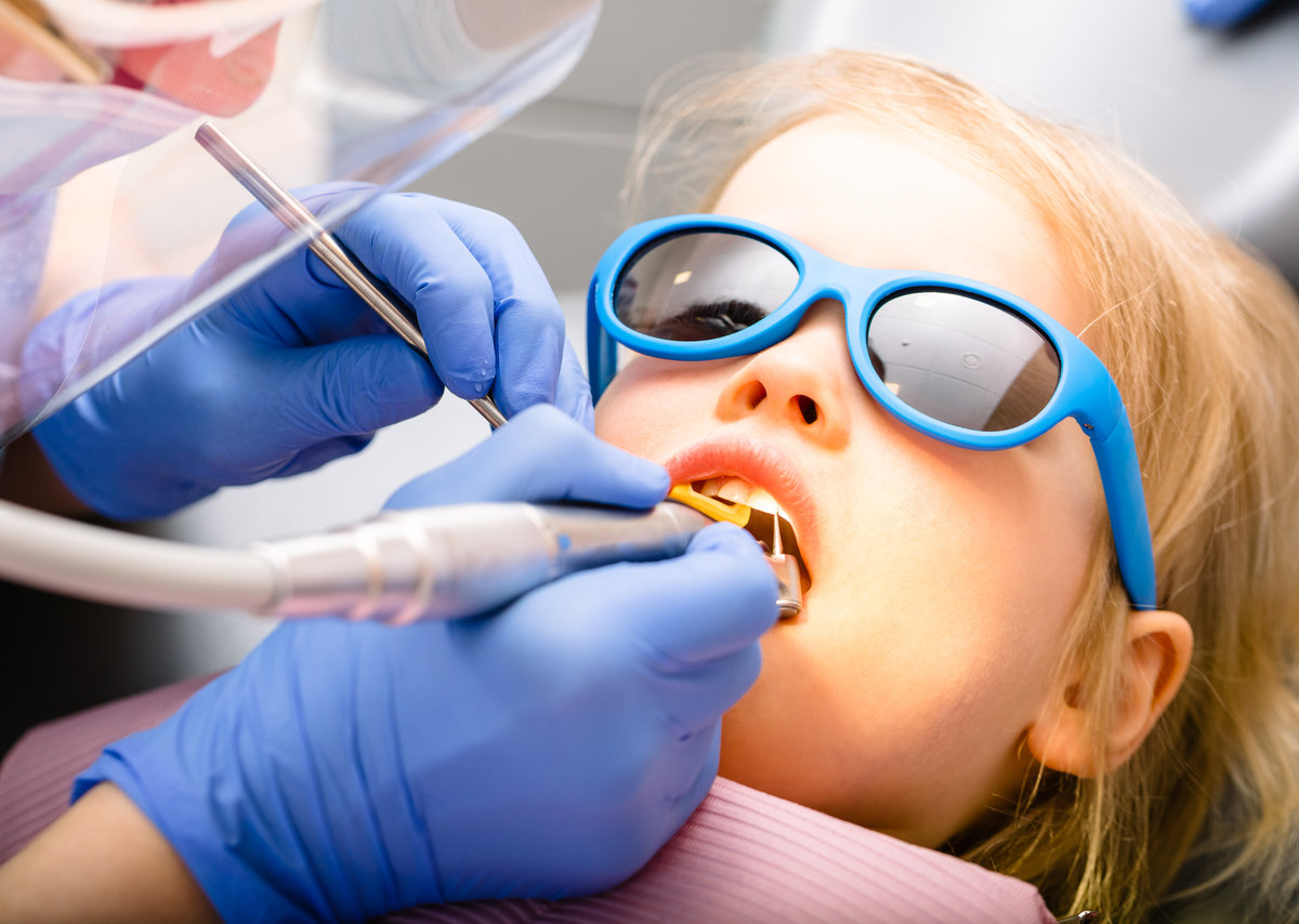 Dentist performing dental filling procedure to a little girl in pediatric dental clinic. Calm child is sitting in a dental chair wearing sunglasses holding mouth open