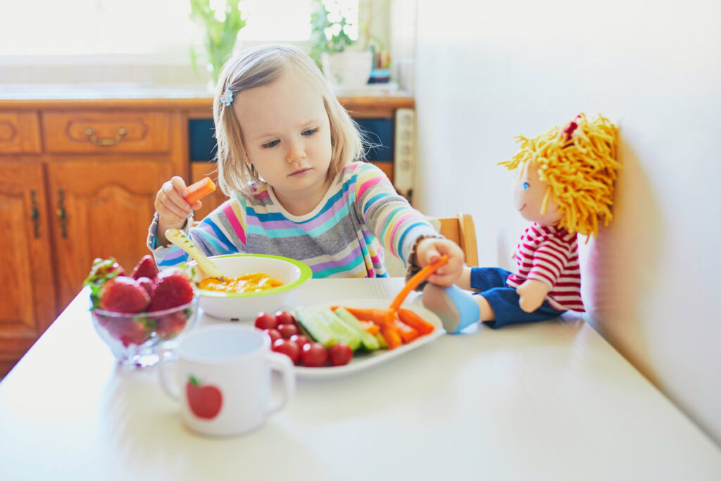 Adorable toddler girl eating fresh fruits and vegetables for lunch. Child feeding doll and teddy bear in the kitchen. Delicious healthy food for kids