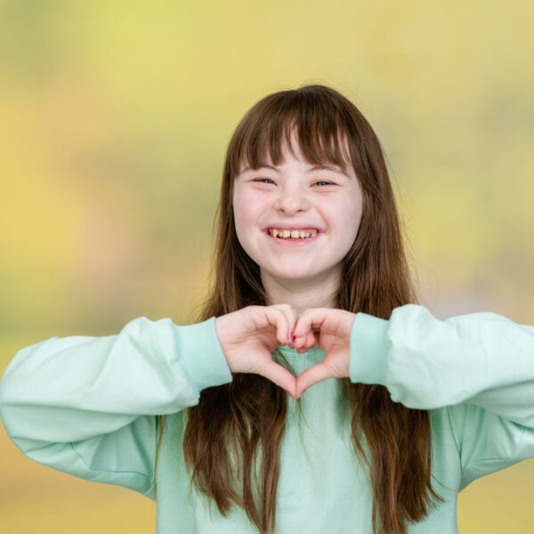 Happy young girl with Downs syndrom shows heart sign at summer park.