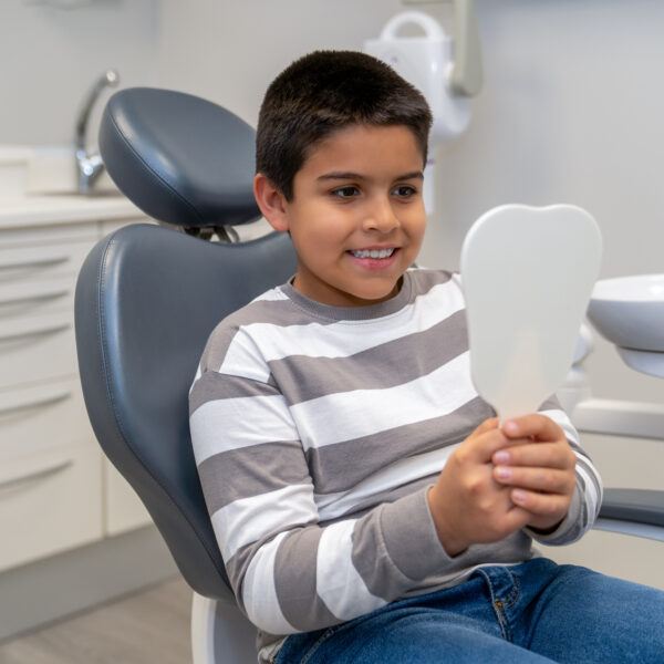 Happy boy sitting in dentist chair, holding a mirror and checking his teeth after dental checkup