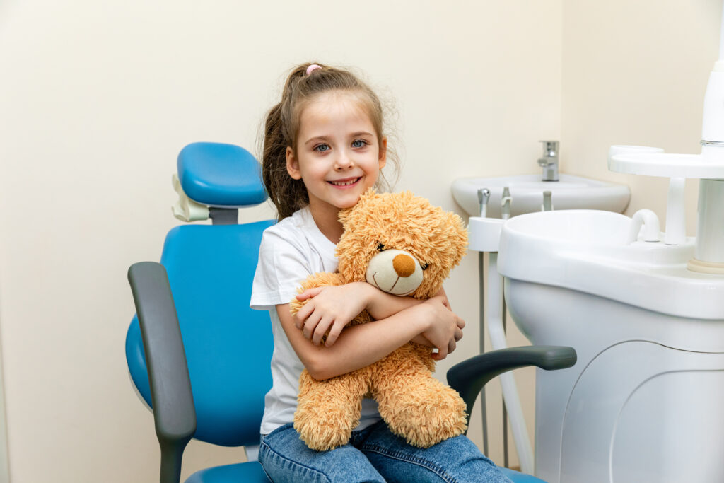Happy little girl having dentist's appointment in modern clinic. Little patient, adorable girl sitting in dentist's chair during appointment in pediatric dentistry clinic and hugging her teddy bear.