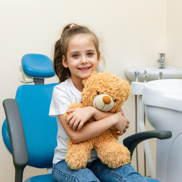 Happy little girl having dentist's appointment in modern clinic. Little patient, adorable girl sitting in dentist's chair during appointment in pediatric dentistry clinic and hugging her teddy bear.