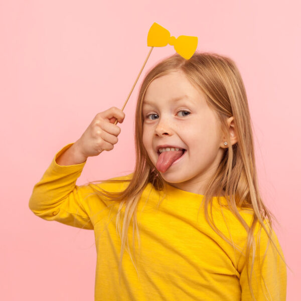 Portrait of playful adorable little ginger girl holding paper bow and sticking out tongue, naughty child having fun, wearing masquerade accessory. indoor studio shot isolated on pink background
