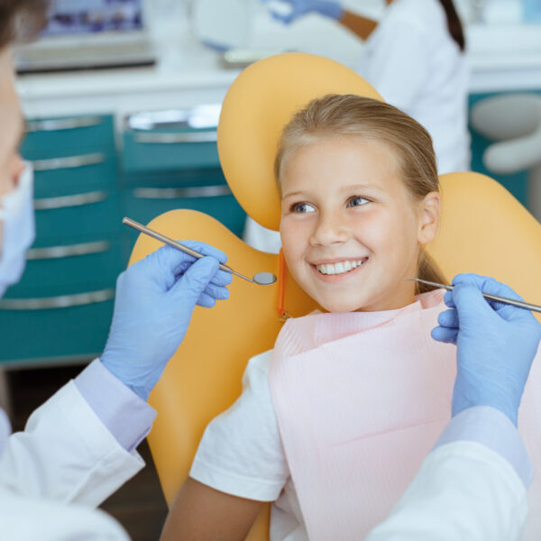 Fearless visit to doctor and dental treatment for child. Smiling girl in medical chair looks at dentist man in white coat, protective mask and rubber gloves with tools in his hands, and not afraid