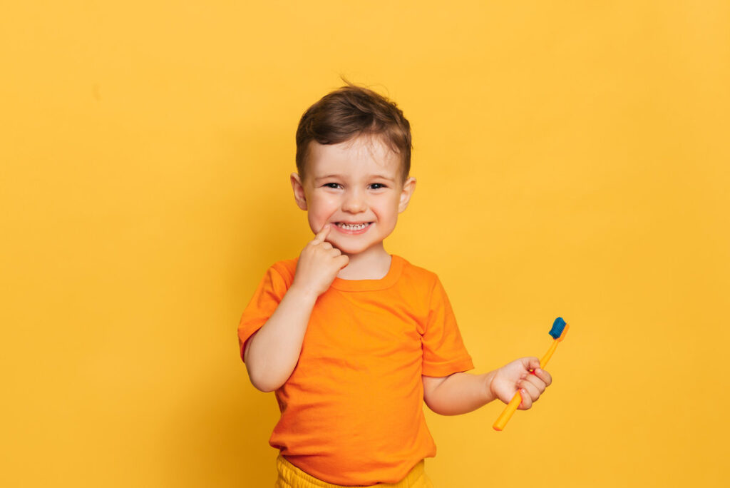 Happy baby toddler boy brushing his teeth with toothbrush on a yellow background. Health care, oral hygiene.