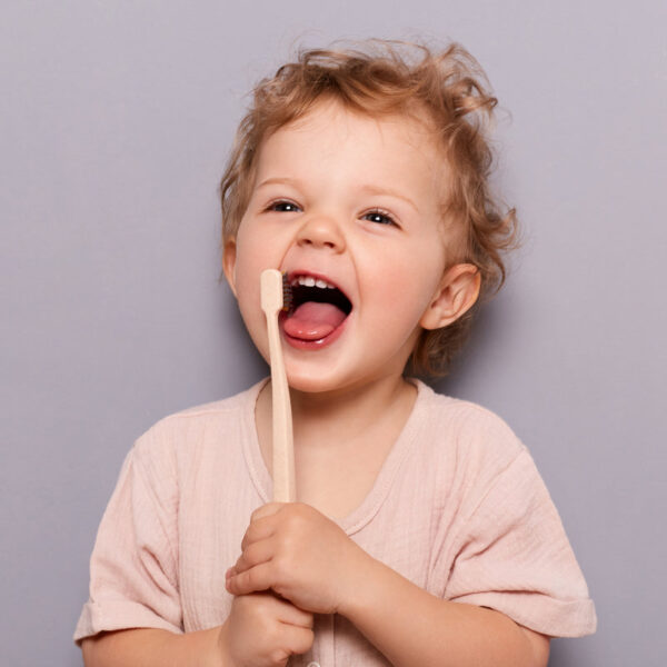 Portrait of laughing happy cheerful infant girl kid with curly blonde hair brushing her teeth, taking care of dental hygiene standing isolated over gray background.