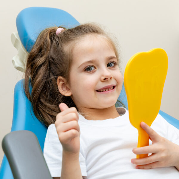Portrait of a cute happy little girl sitting on a chair in a dental office, showing thumbs up after a dental examination while holding mirror in her hand.