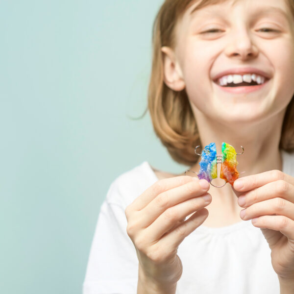 Smiling child holds rainbow orthodontic retainer on pastel background with copy space for dental clinic banner and kids care promotion.