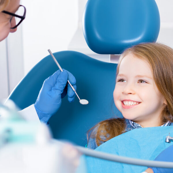 Little cute smiling girl is sitting in dental chair and opening mouth in clinic, office. Doctor is preparing for examination of child teeth with tools, instruments. Visiting dentist with children.