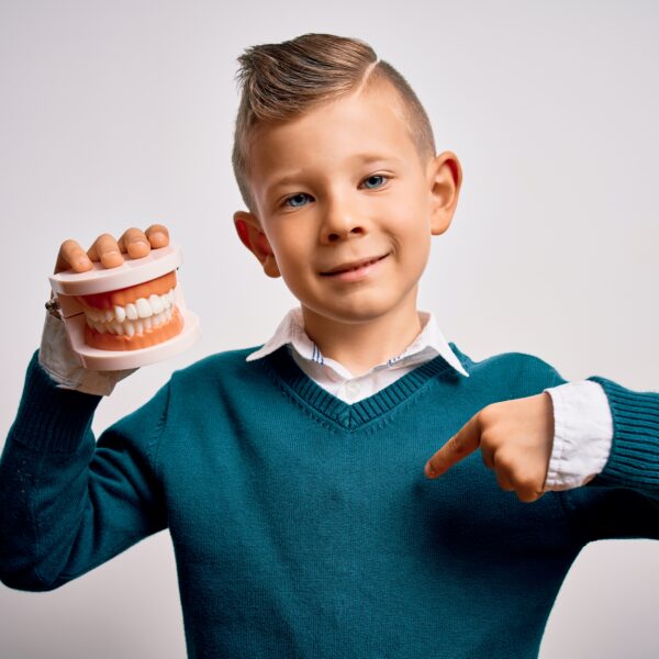 Young little caucasian kid holding dental prosthesis teeth denture over isolated background with surprise face pointing finger to himself